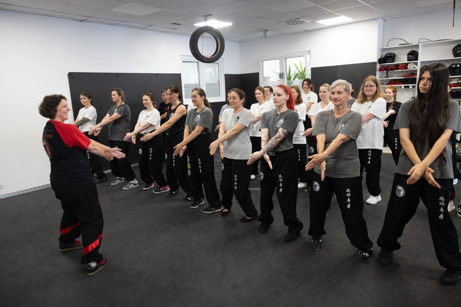 Eine große Gruppe Erwachsener steht in einer Reihe im Trainingsraum und übt eine Handbewegung. Die Trainerin steht ihnen zugewandt gegenüber. Die Teilnehmerinnen und Teilnehmer sind unterschiedlichen Alters, von Anfang zwanzig bis über sechzig.